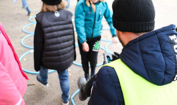 Utomhus, en lärare i reflexväst har ryggen mot kameran, två elever på skolgården där det är spraymålat ringar på asfalten för en lek. 